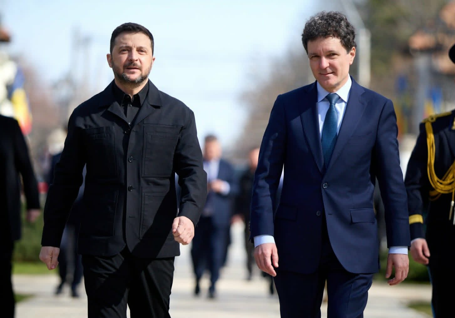 Two men in formal attire walk outside side by side, one in a black jacket and the other in a navy suit, with a blurred background behind them.