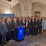 Group of suited politicians and staff pose at a press event around a blue podium with the PNL logo in a grand room.