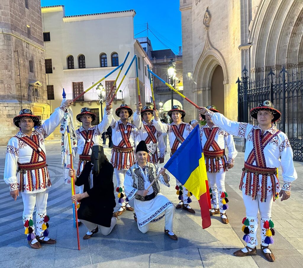 Group of men in ornate Eastern European folk costumes holding colorful poles, with a Ukrainian flag, in a town square near a church.