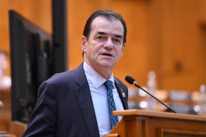 Man in a dark suit and blue patterned tie speaks at a wooden podium with a microphone in a formal conference setting.