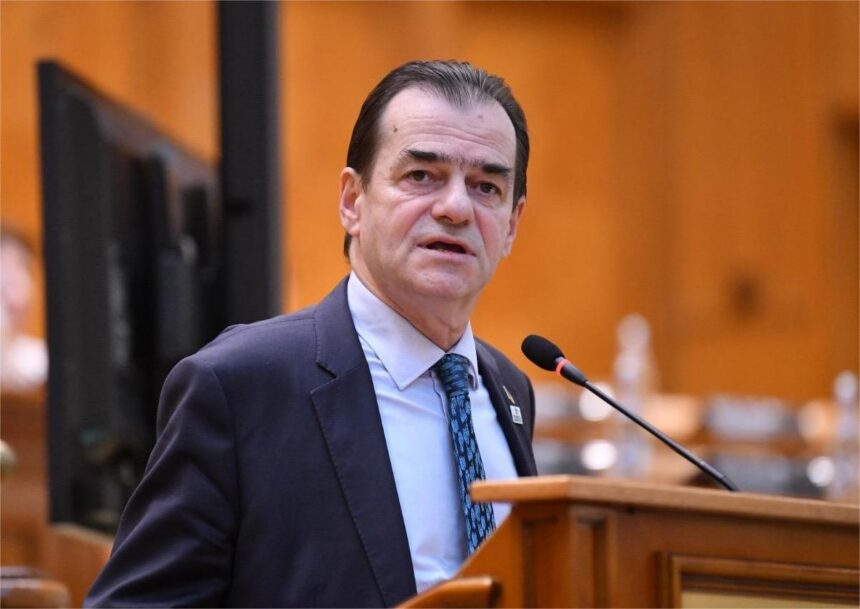 Man in a dark suit and blue patterned tie speaks at a wooden podium with a microphone in a formal conference setting.