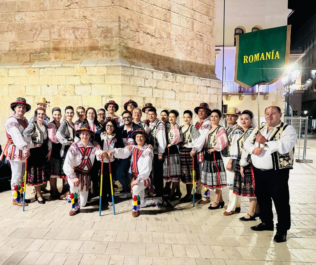 Group of people in colorful Romanian folk costumes posing for a night photo, one man holding a green 'ROMANIA' banner near a stone wall.