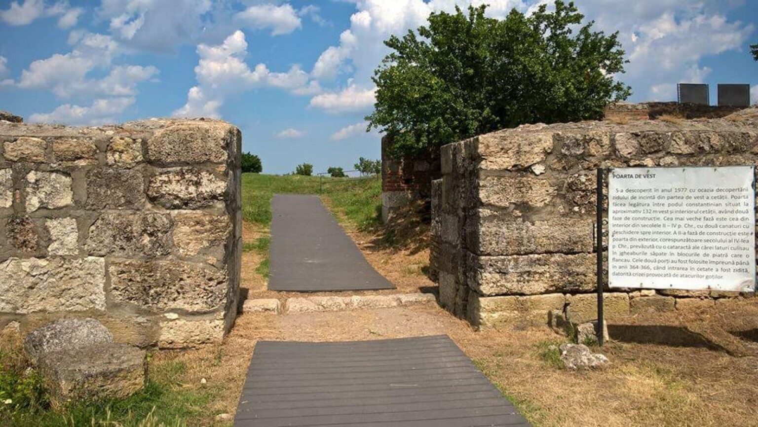 Stone gateway with a paved path through a grassy area under a blue sky; a signboard with text on the right.