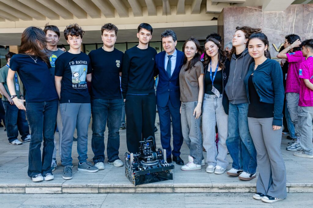 Group of students posing with a suited man and a small robotics platform in front of a building exterior.