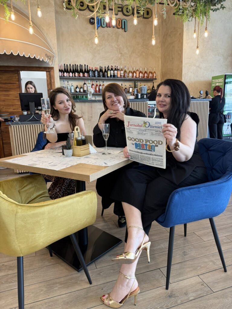 Three women seated at a restaurant table raising champagne glasses, smiling at the camera with a newspaper on the table and drinks behind them.