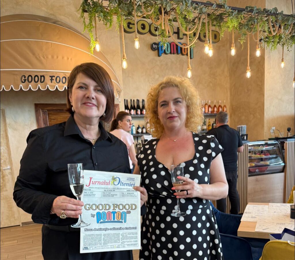 Two smiling women pose in a cozy cafe; one wears black and holds a newspaper and a champagne flute, the other in a polka-dot dress with a glass in hand, near a bar backdrop.