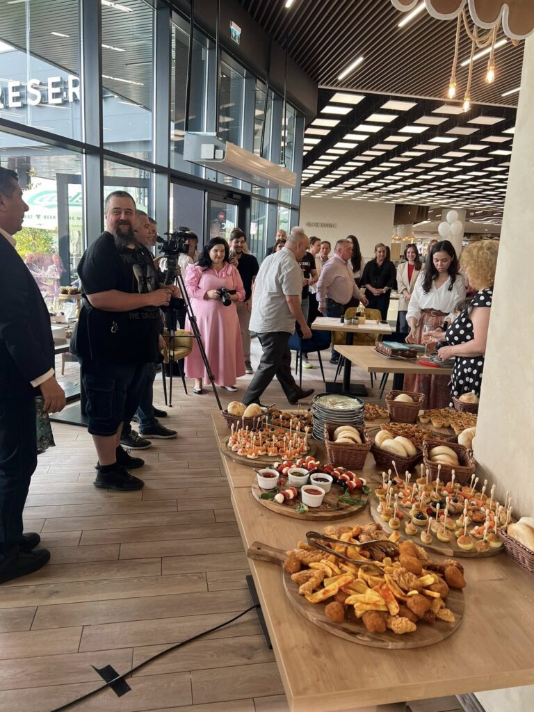 A group of people at a catered event in a modern glass-walled venue, with a photographer filming and tables full of appetizers and desserts in the foreground.