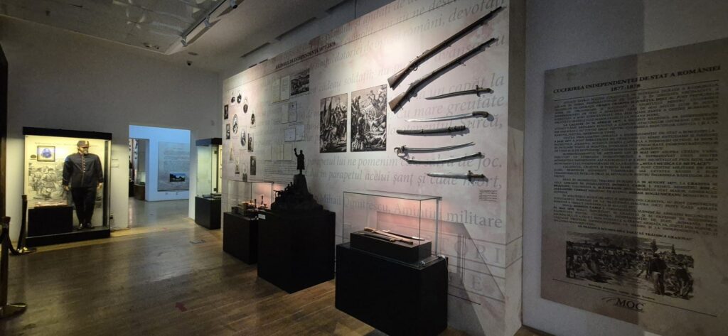 Museum gallery showing historical weapons mounted on wall and glass display cases, with Romanian text panels and a statue pedestal in the center.