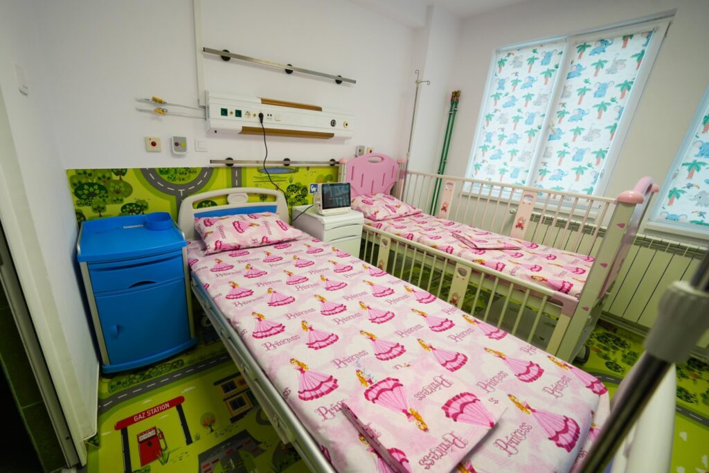 Pediatric hospital room with two pink princess beds, medical panels, and elephant-themed window blinds on a bright wall.