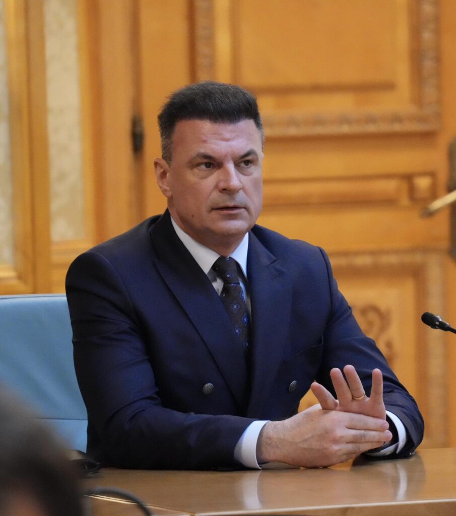 Man in a dark suit and tie seated at a wooden conference table, hands resting on the table, in a formal setting.