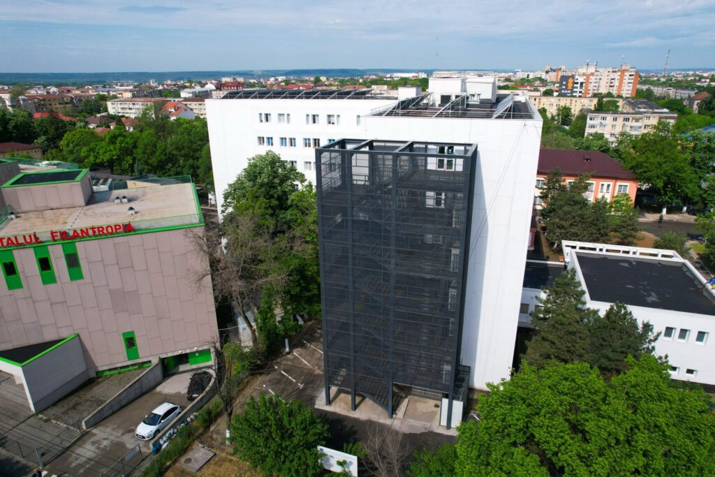 Aerial view of a tall glass-fronted building next to a white rectangular office block in a green urban area, with trees and streets nearby.