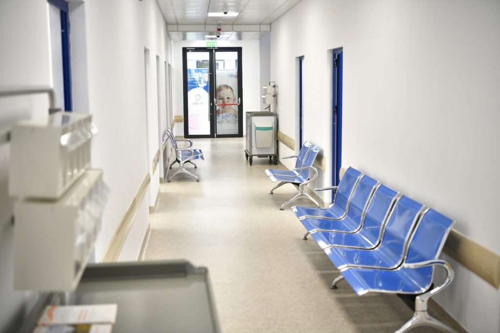 Hospital corridor with blue chairs along the right wall and glass double doors at the far end showing a poster.