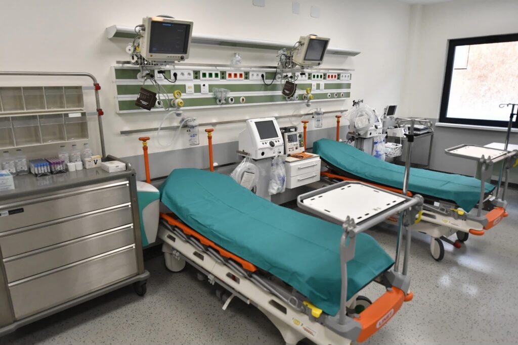Hospital patient care area with two wheeled stretchers covered in teal sheets and multiple monitors mounted on the wall above medical gas panels and cables.