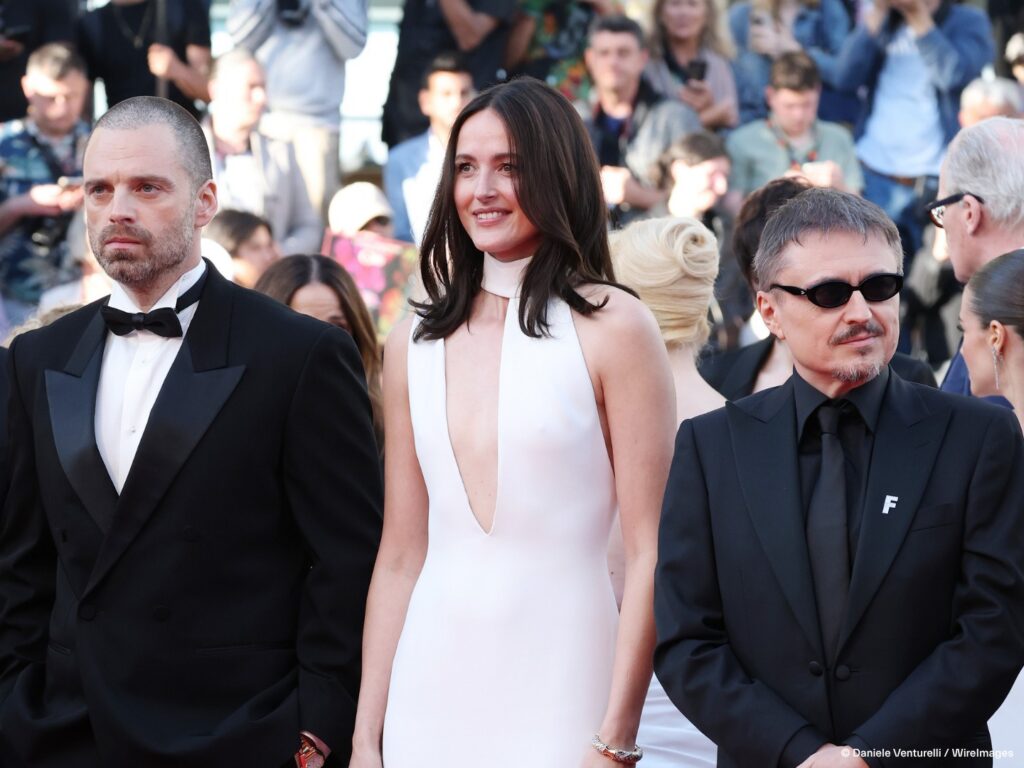 Three celebrities in formal attire on a red carpet with photographers in the background.
