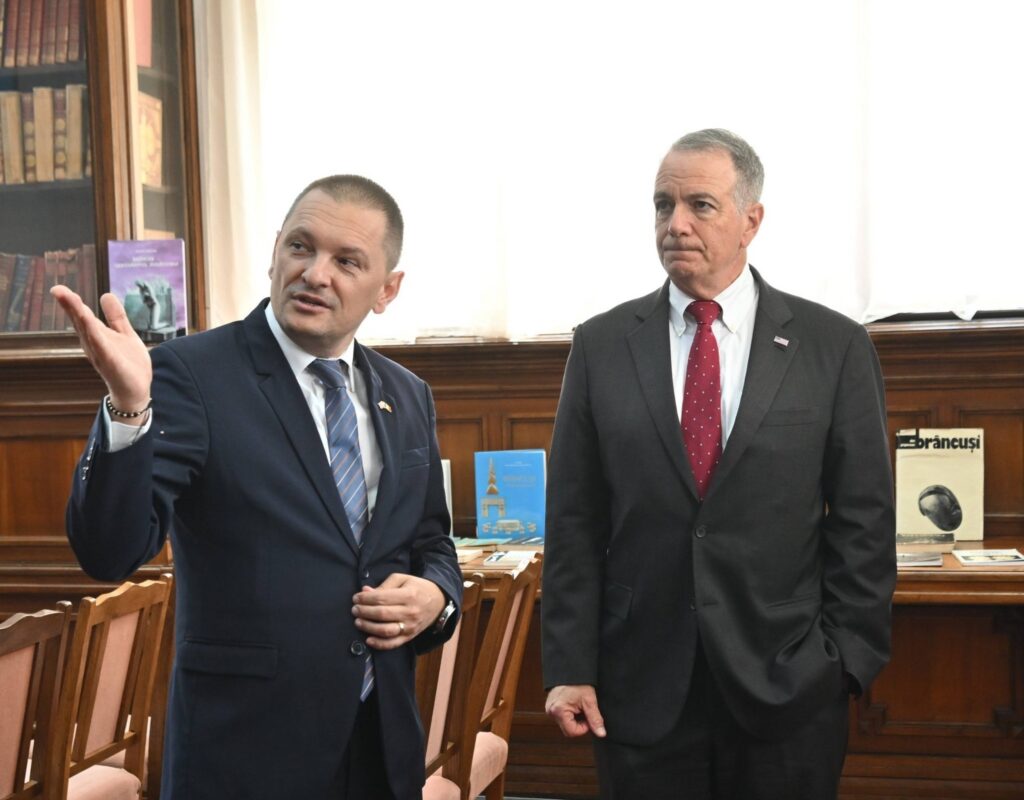 Two men in suits stand in a wood-paneled room with bookshelves; the left man gestures as he speaks.