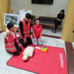Group of young trainees in pink vests practicing CPR on a mannequin on a red mat in a public hallway.