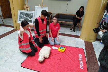 Group of young trainees in pink vests practicing CPR on a mannequin on a red mat in a public hallway.