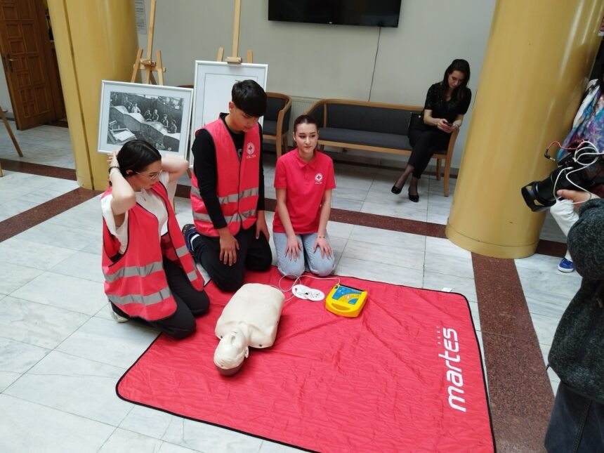 Group of young trainees in pink vests practicing CPR on a mannequin on a red mat in a public hallway.