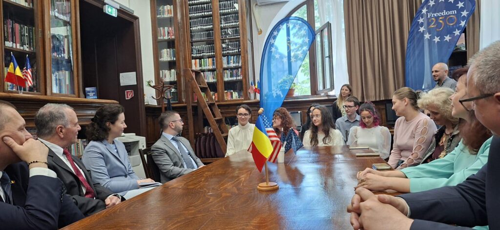 Group of people seated around a wooden table in a library during a formal event, with Romanian and American flags on the table and a blue 'Freedom 250' banner nearby.