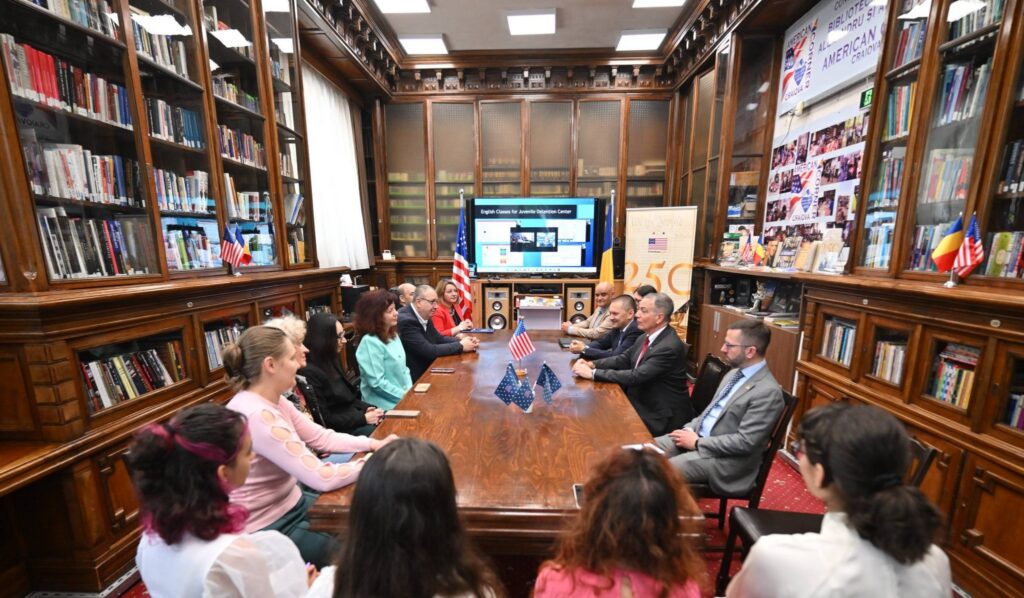 Group of people seated around a long wooden conference table in a historic library, with bookcases lining the room and small flags on the table.
