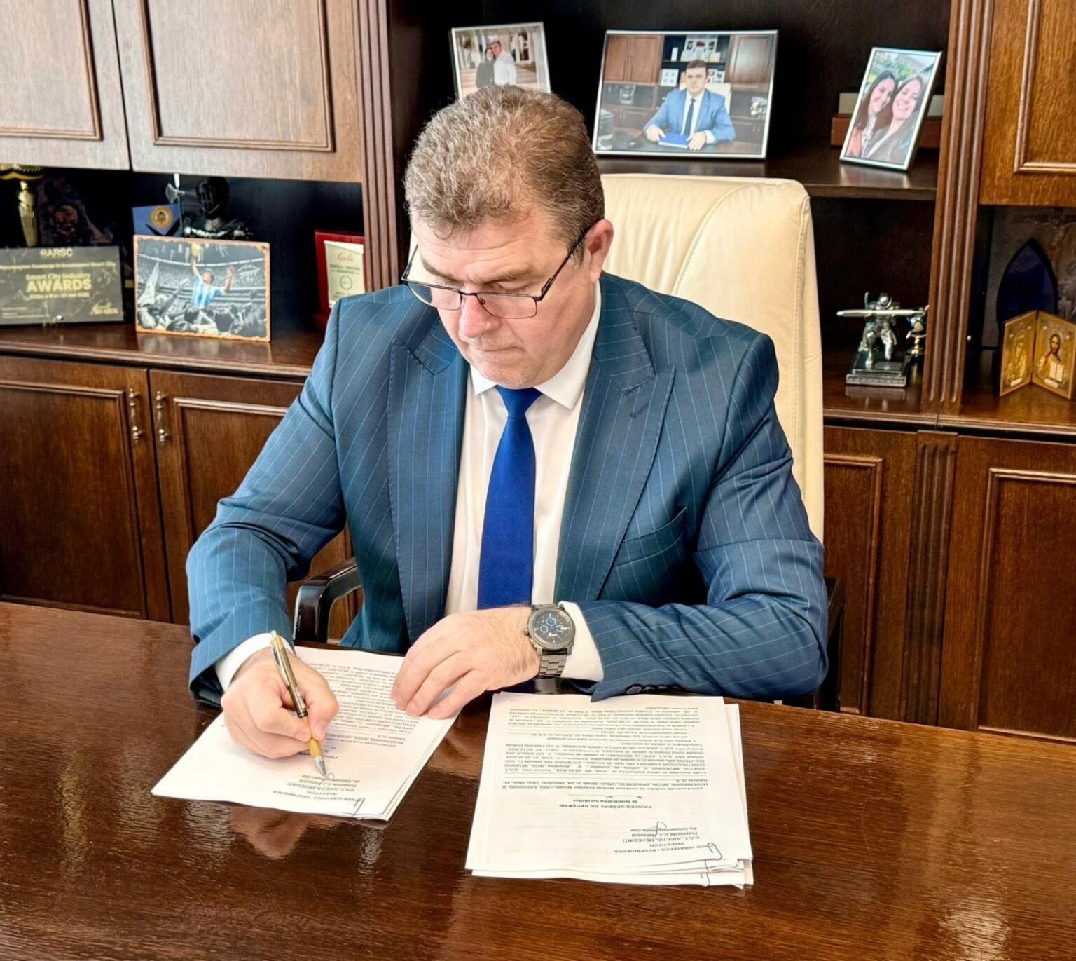 Man in a blue pinstripe suit signs documents at a wooden desk in an office with framed photos and awards behind him.