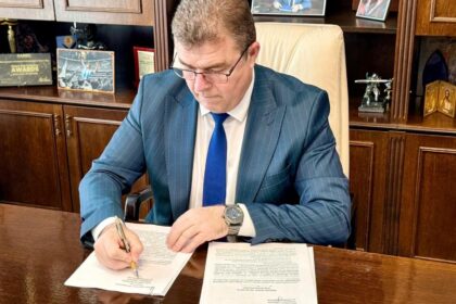 Man in a blue pinstripe suit signs documents at a wooden desk in an office with framed photos and awards behind him.