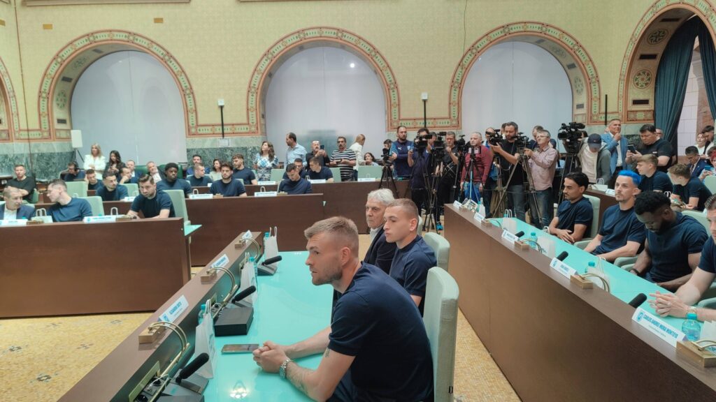 Conference room filled with attendees seated at turquoise desks, photographers and reporters with cameras along the back wall.