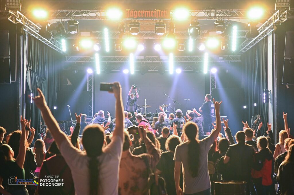 Crowd with raised hands watching a live concert on a brightly lit stage, band performing under multiple spotlights with a Jägermeister banner above.