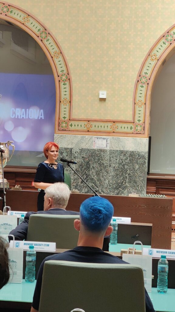 Woman with short red hair speaking at a podium in a formal meeting room with ornate arches behind her.