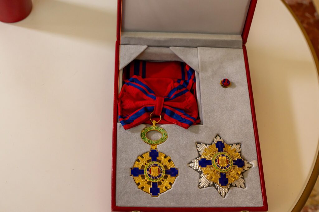 - Jurnalul Olteniei – Știri din Craiova și Oltenia Red presentation box containing two ornate gold and blue cross medals with red-and-blue ribbons, resting on a gray padded insert.