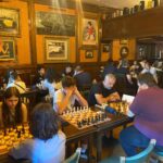 Group of people playing chess in a cozy vintage-style cafe with wood-paneled walls and framed art behind them