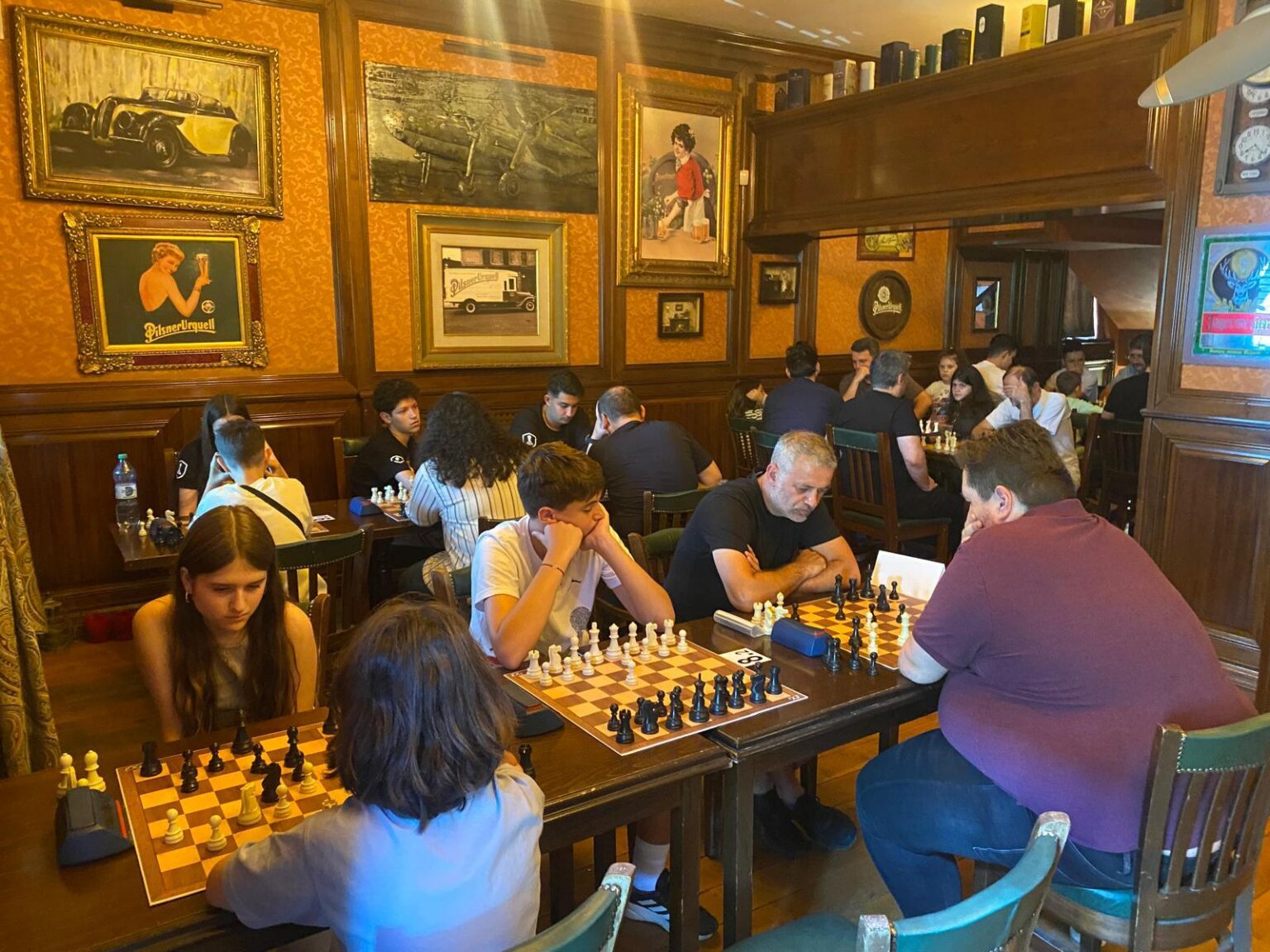 Cupa Firmelor la Șah - Jurnalul Olteniei – Știri din Craiova și Oltenia Group of people playing chess in a cozy vintage-style cafe with wood-paneled walls and framed art behind them