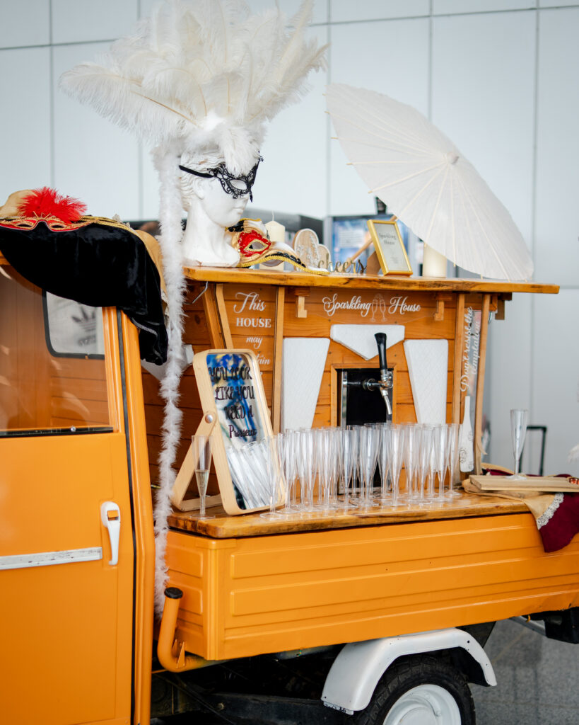 Champagne bar on an orange vehicle with a white feathered masquerade headdress, black mask, and a white parasol, lined with flutes.
