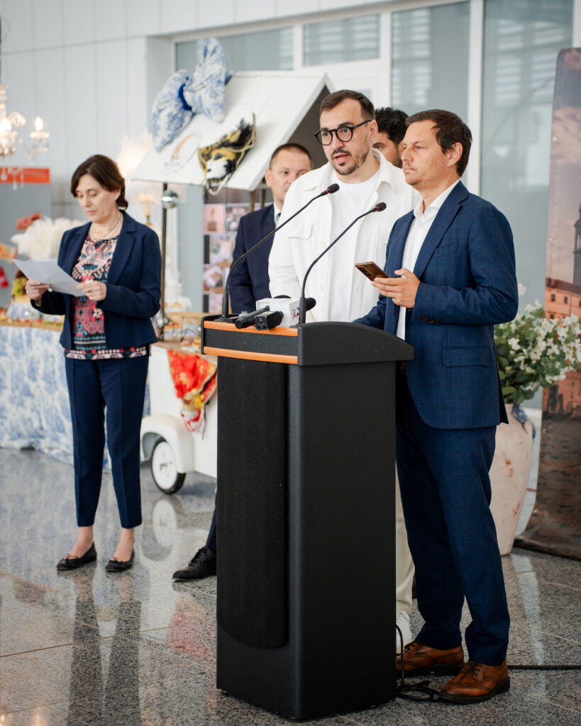 Speaker at a podium with two microphones, flanked by colleagues during an indoor press event.