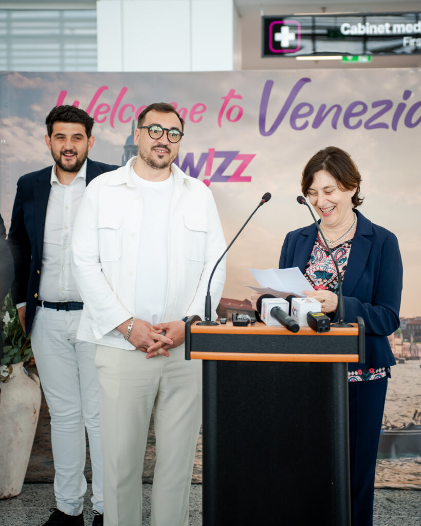 Three people at a press event stand behind a podium with multiple microphones, in front of a 'Welcome to Venezi' banner.