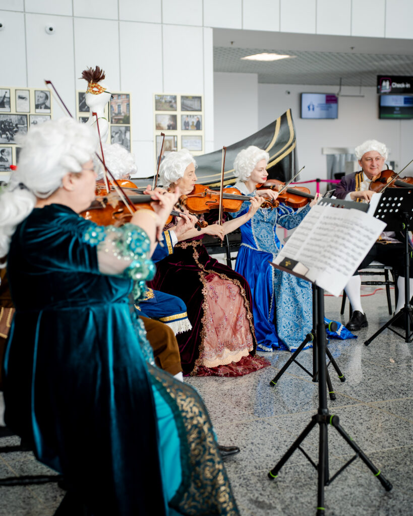 Group of senior musicians in ornate 18th-century costumes playing violins in a bright indoor space, with music stands and sheet music nearby