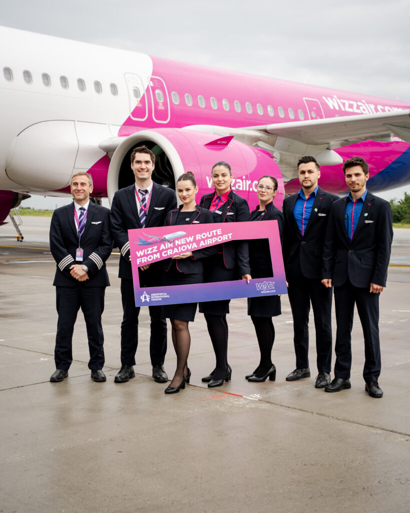 Group of airline staff in navy uniforms posing on the tarmac in front of a pink Wizz Air plane, holding a bright pink sign promoting a new route from Craiova Airport.