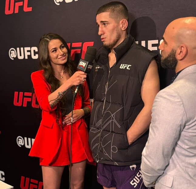 Female reporter in a red dress interviews a UFC fighter wearing a black vest; another man in a light suit stands nearby, backstage with UFC branding.