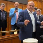 Man in a navy suit gestures toward two fundraising buckets on a desk in a wood-paneled council chamber, flanked by colleagues looking on.