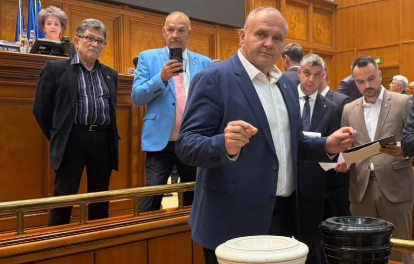 Man in a navy suit gestures toward two fundraising buckets on a desk in a wood-paneled council chamber, flanked by colleagues looking on.