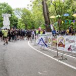 Group of runners gathered at a park start line with sponsor banners and colorful bunting among green trees in the background.
