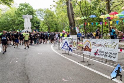 Group of runners gathered at a park start line with sponsor banners and colorful bunting among green trees in the background.