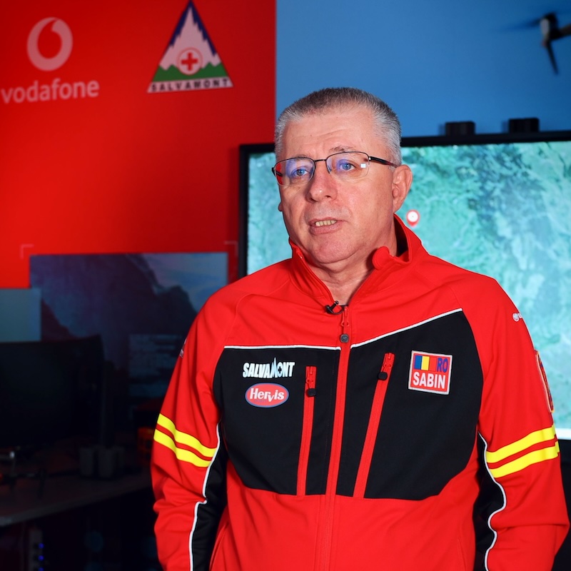 Man in a red and black jacket with sponsor logos speaks on camera in a tech demo room, Vodafone and mountain logos visible in the background