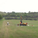 Drilling rig on a grassy rural field with two workers nearby, performing a drilling operation in a wide-open landscape.