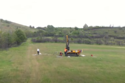 Drilling rig on a grassy rural field with two workers nearby, performing a drilling operation in a wide-open landscape.