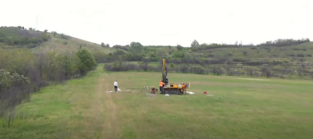 Drilling rig on a grassy rural field with two workers nearby, performing a drilling operation in a wide-open landscape.