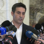 Man in a dark suit speaks to a cluster of microphones held by reporters indoors, during a press event.