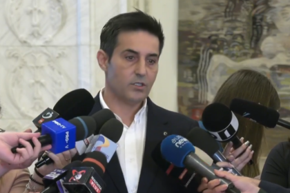 Man in a dark suit speaks to a cluster of microphones held by reporters indoors, during a press event.