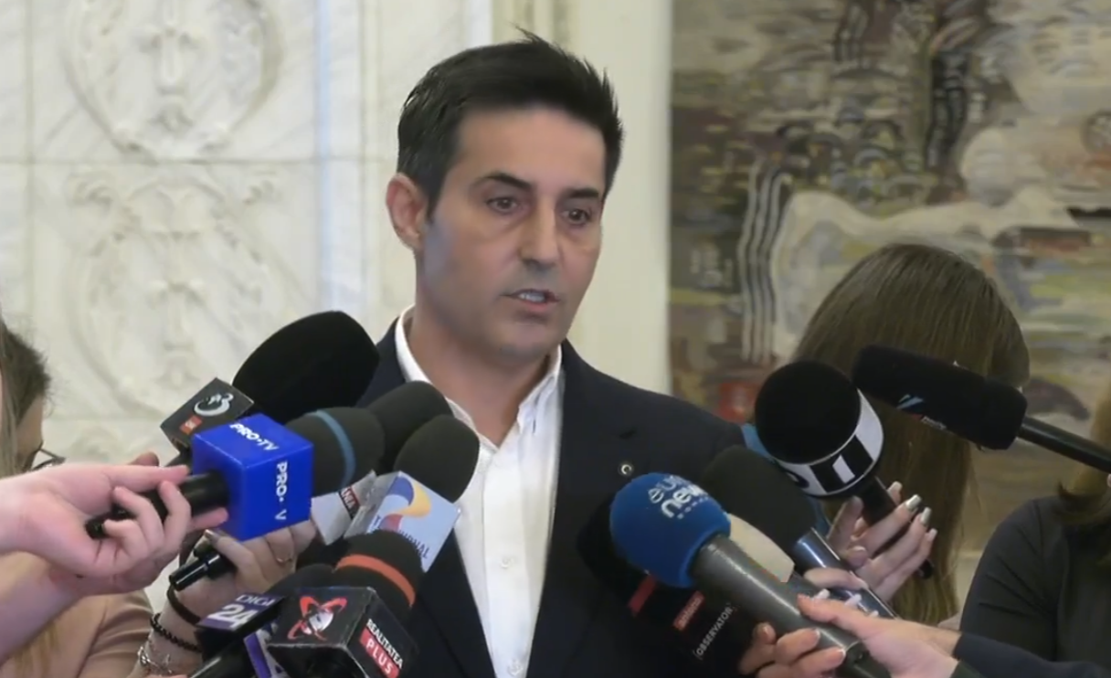 Man in a dark suit speaks to a cluster of microphones held by reporters indoors, during a press event.