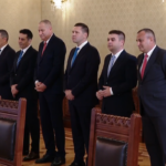 Six men in dark suits stand in a row in a formal room, hands clasped, facing forward near ornate wooden chairs and patterned wallpaper.